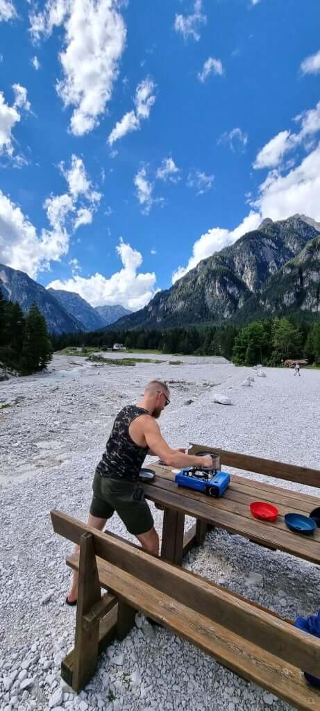 Person cooking by the river with mountain backdrop